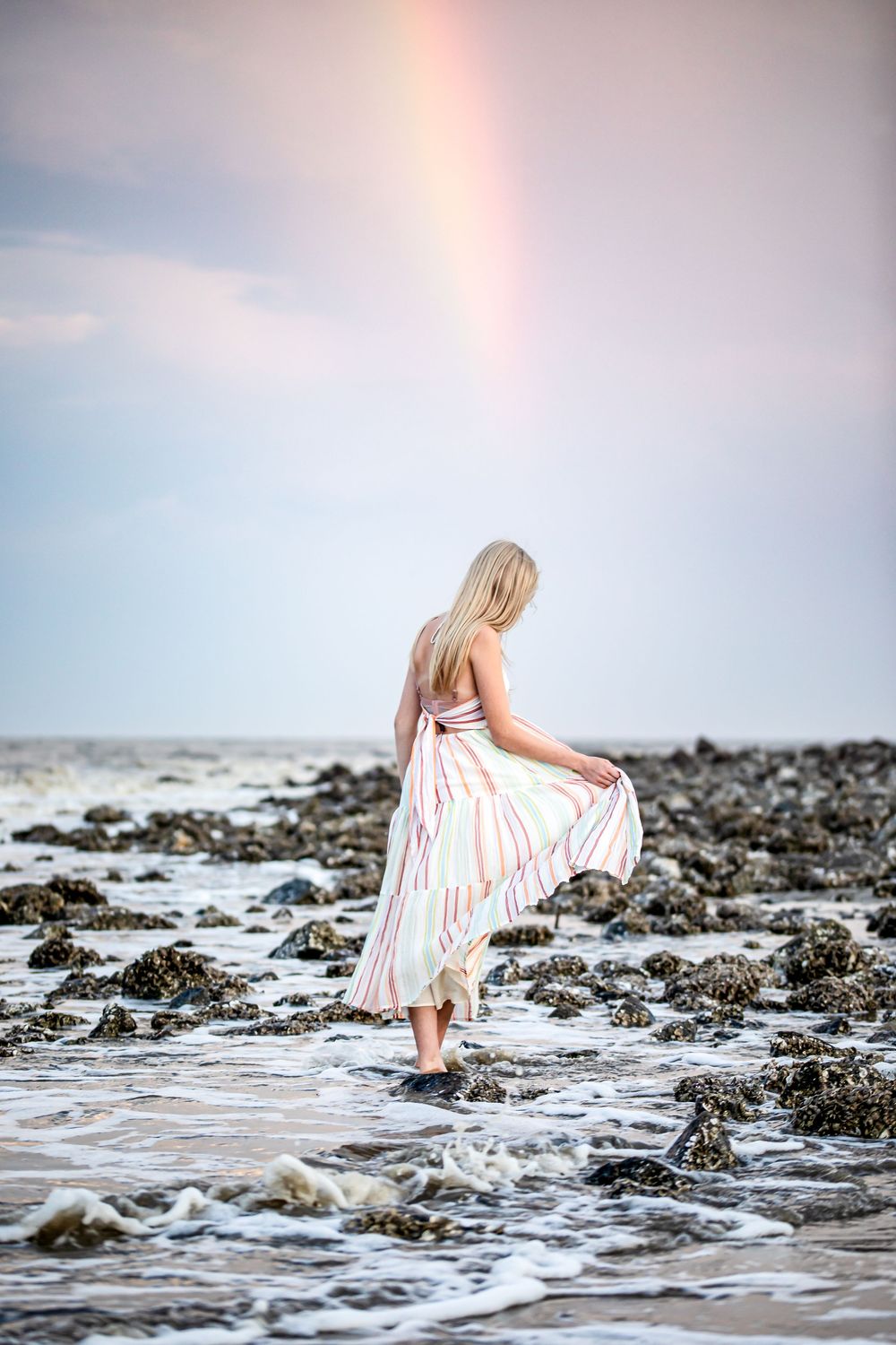 Woman in white dress walks along rocky shoreline as waves crash against the coast during sunset.