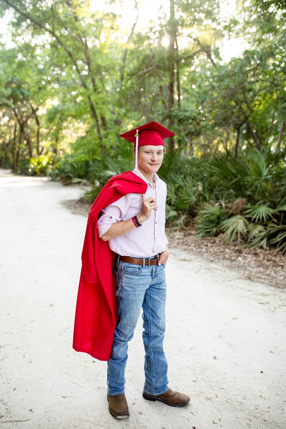 Young man in red graduation cap and gown poses on sandy path surrounded by lush green trees.