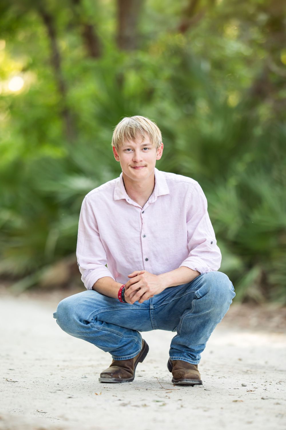 Smiling blonde teenage boy in pink shirt and jeans crouching on sandy path with lush green trees in background.
