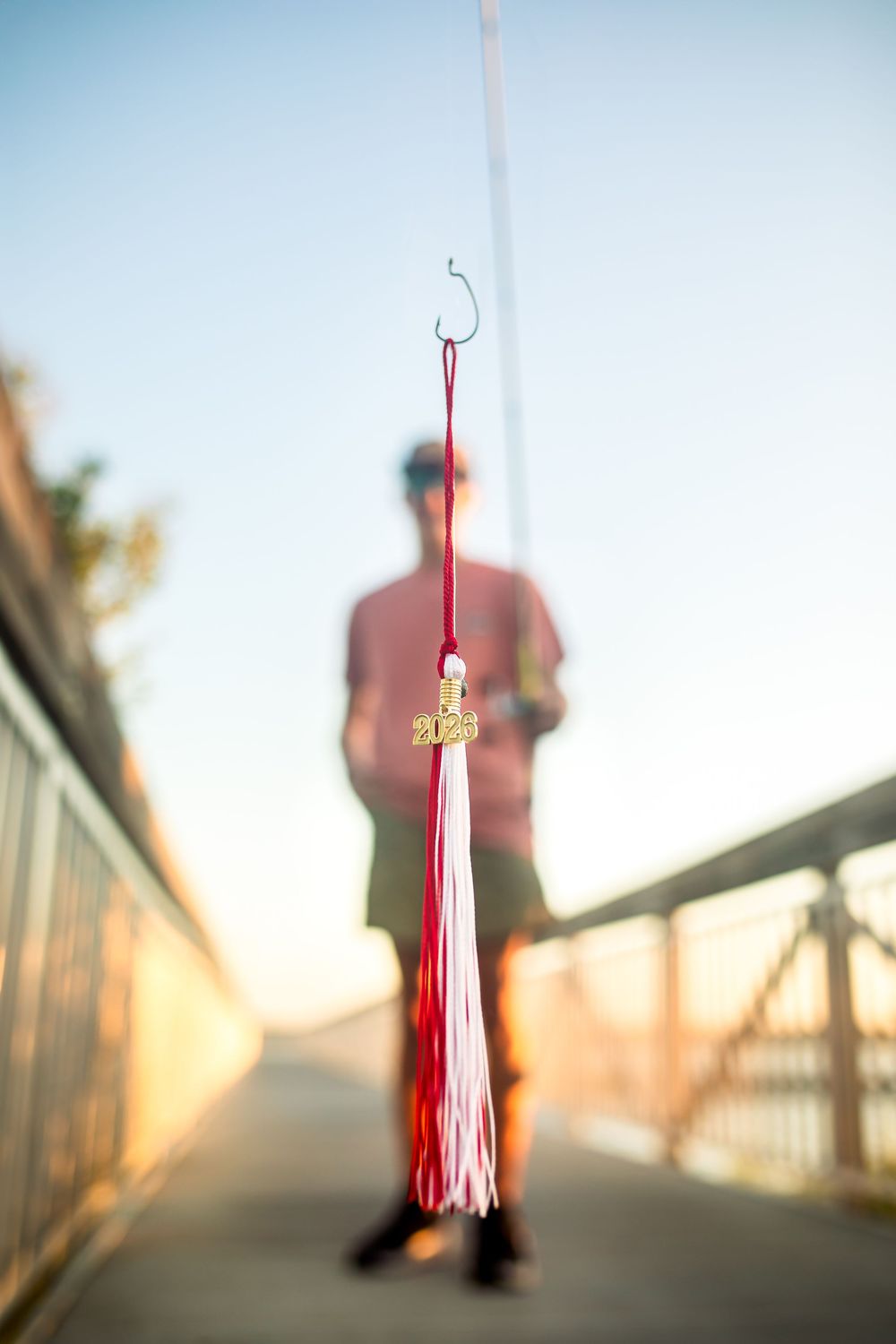 A red and white 2023 graduation tassel hangs from a fishing hook with a blurred graduate standing on a bridge.