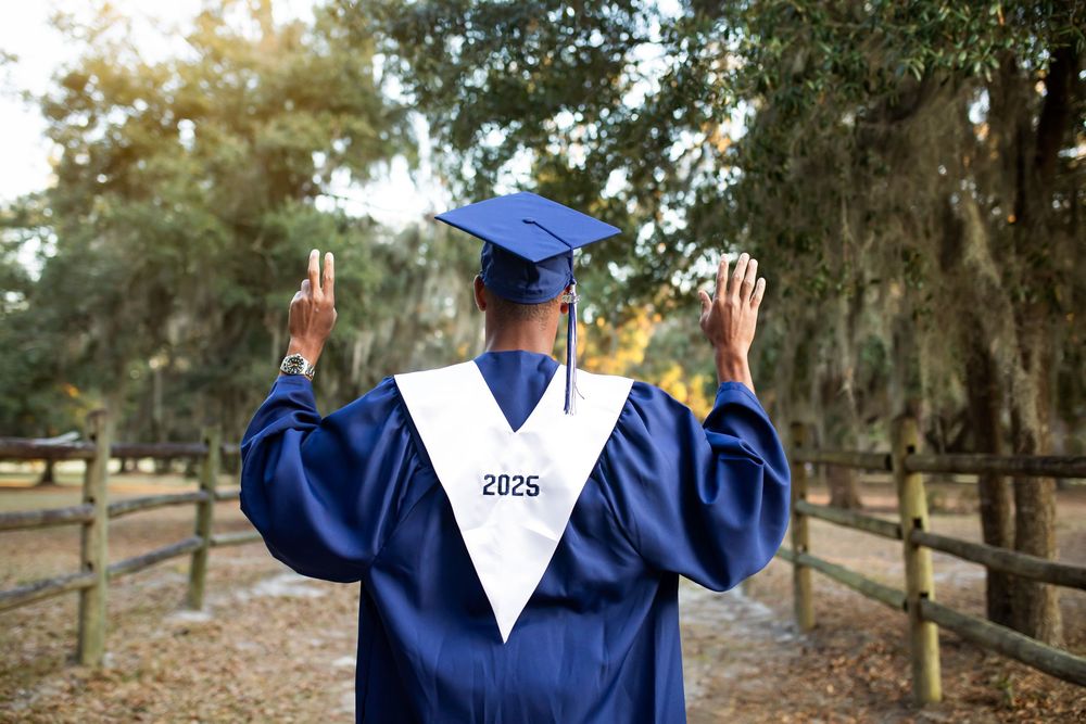 A person in a navy graduation gown and cap poses for celebratory graduation photos at a tree-lined path.