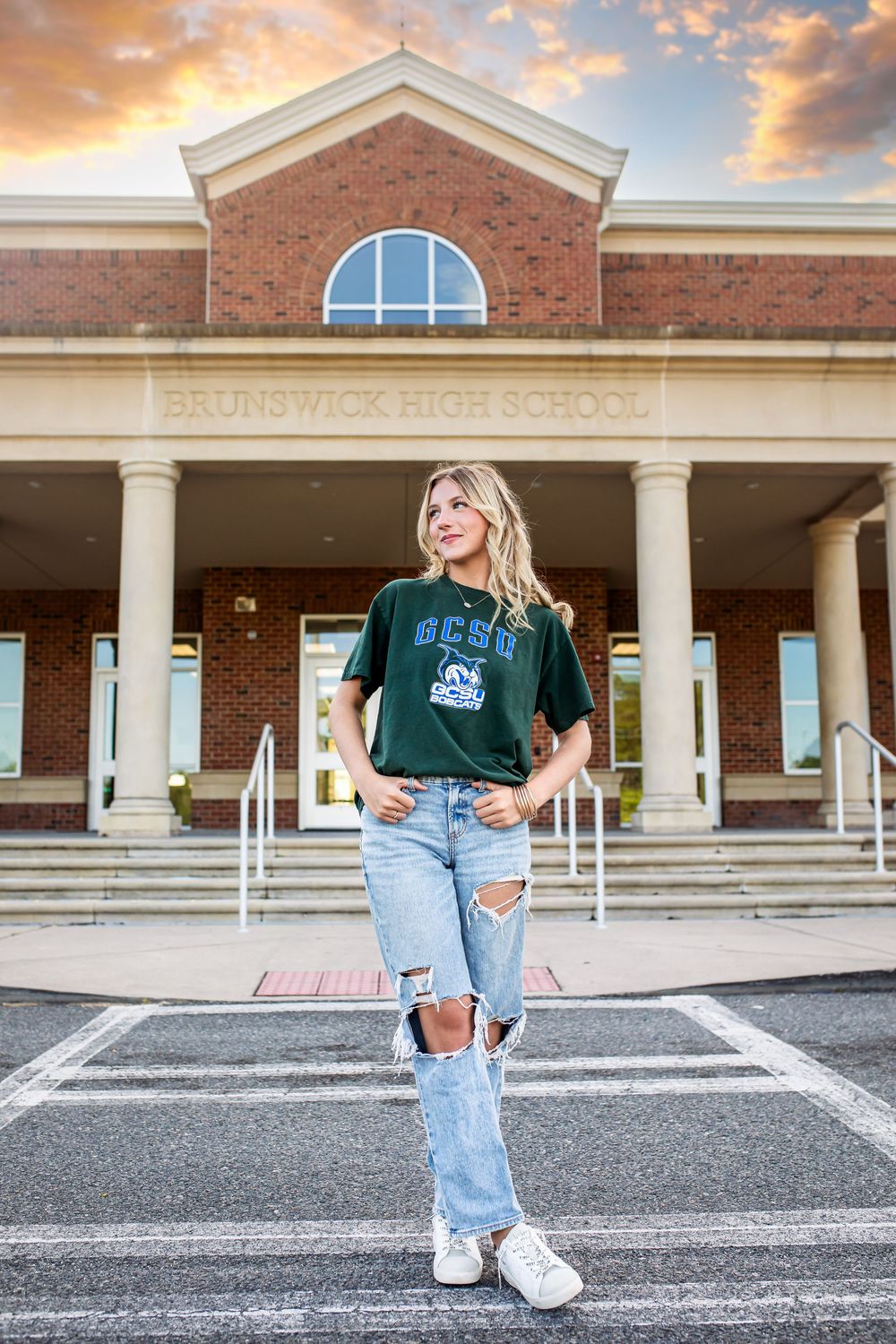 Student in green university t-shirt and ripped jeans poses in front of brick campus building during golden sunset.