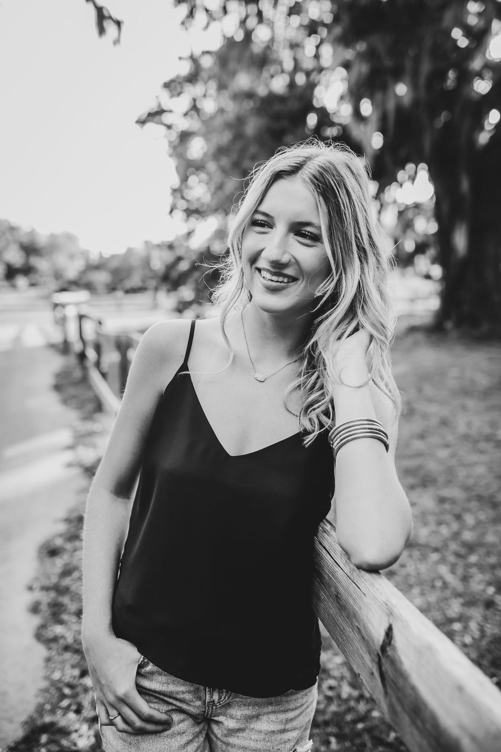 Black and white portrait of someone in a tank top smiling outdoors by a wooden fence on a sunny day.
