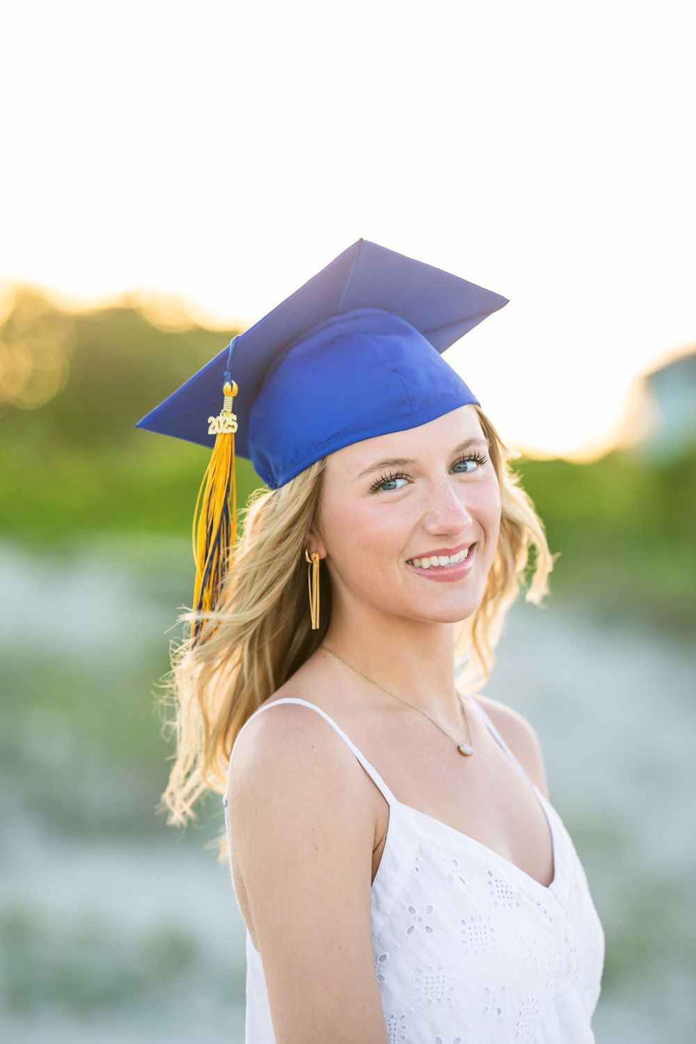 A graduate in a blue cap and white dress smiles brightly during outdoor graduation photos at sunset.