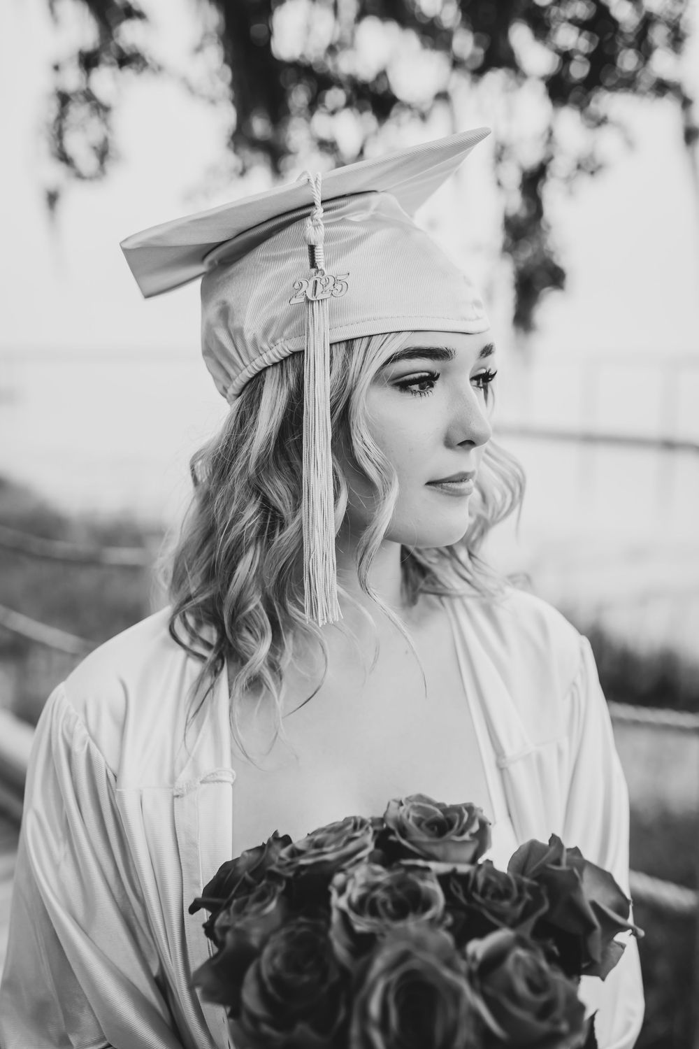 Black and white graduation photo with person in cap and gown holding bouquet of roses while looking to the side.