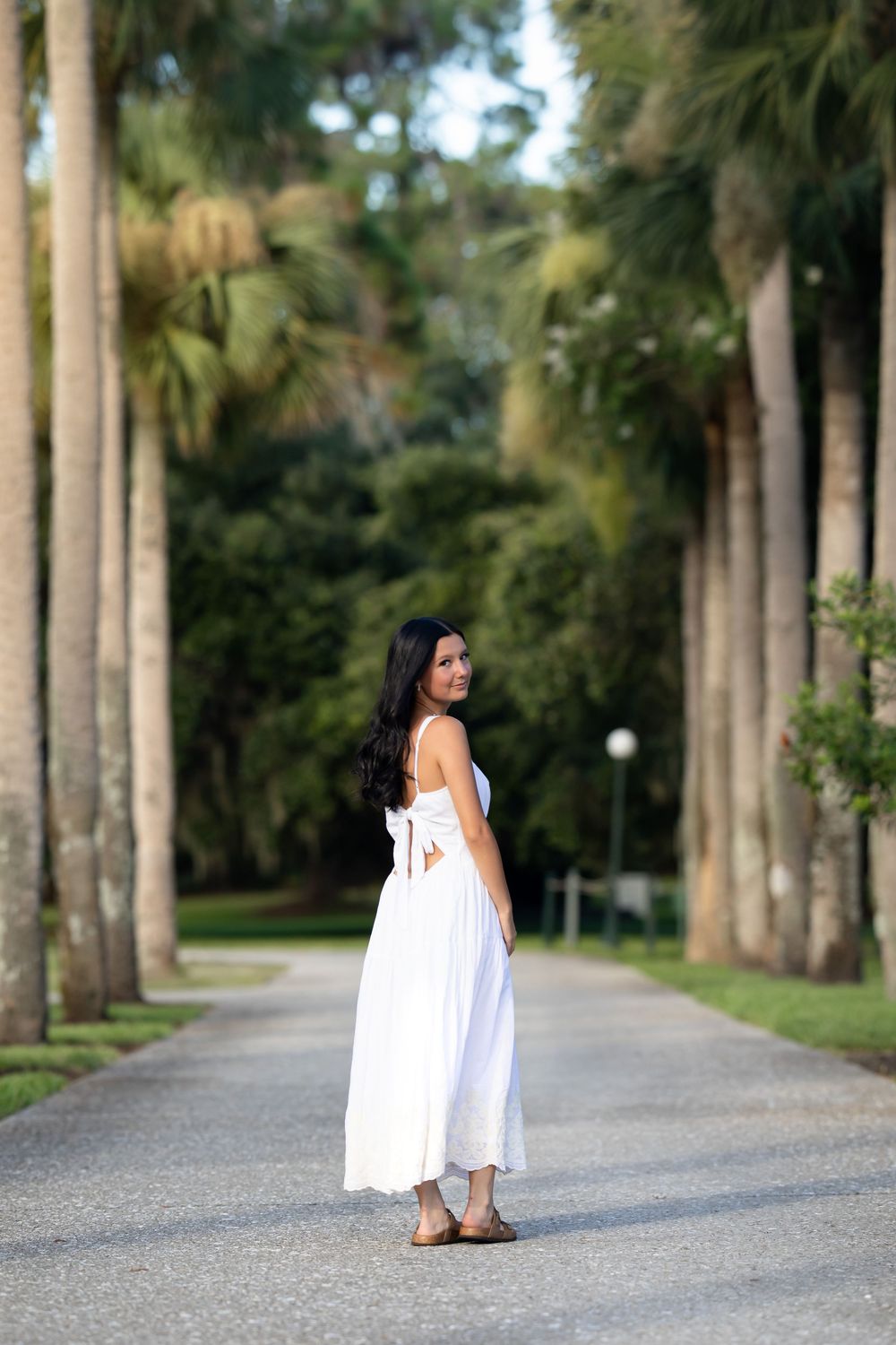 A person in a flowing white dress poses on a scenic palm tree-lined path in a tropical garden setting.