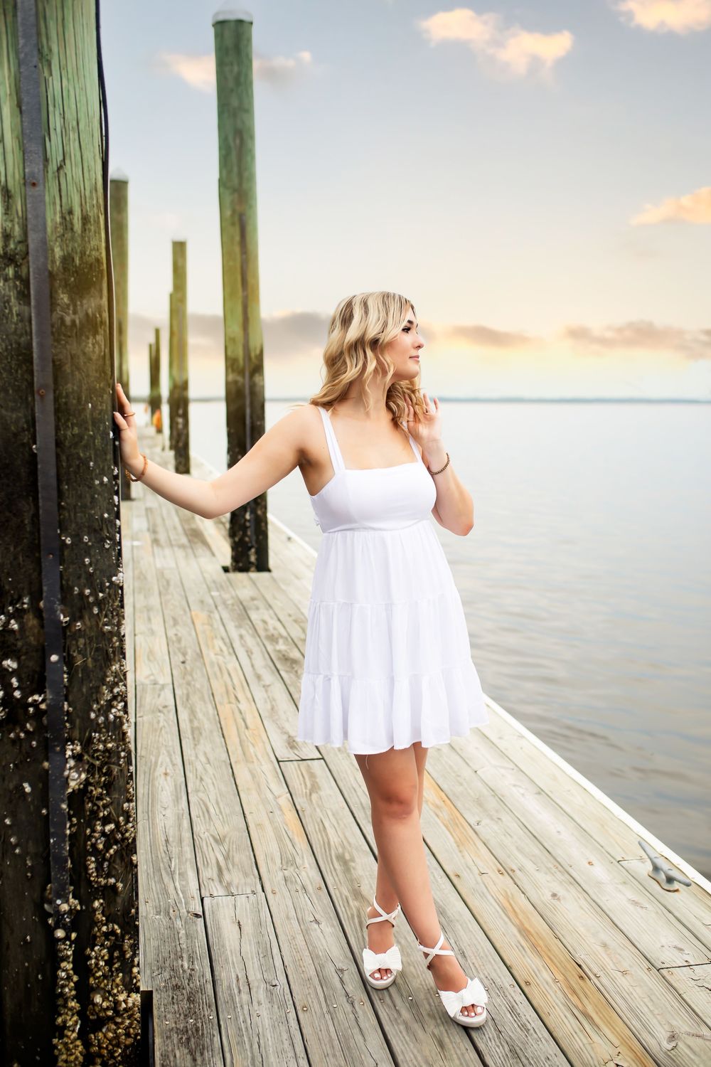White sundress on wooden pier at sunset with tall green posts and calm water views.