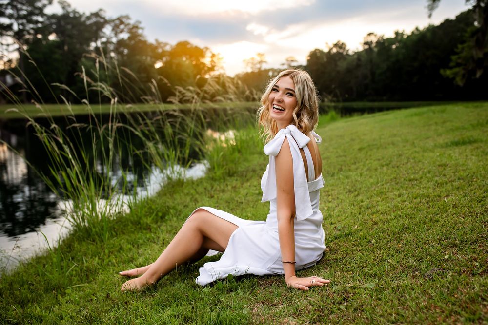 Person in white dress sitting on grassy bank beside pond at sunset with golden light filtering through trees.