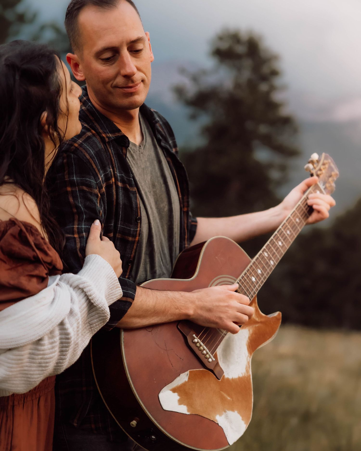 A couple stands close together in a field at sunset while holding an acoustic guitar and sharing an intimate moment.