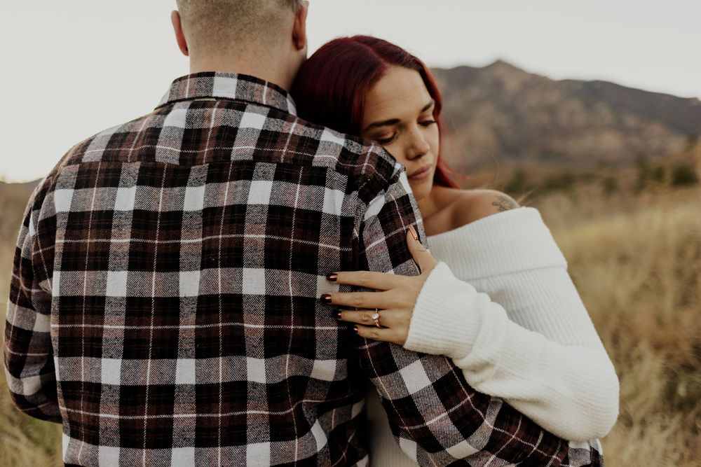 A couple in plaid shirts embraces against scenic mountain landscape during golden hour.
