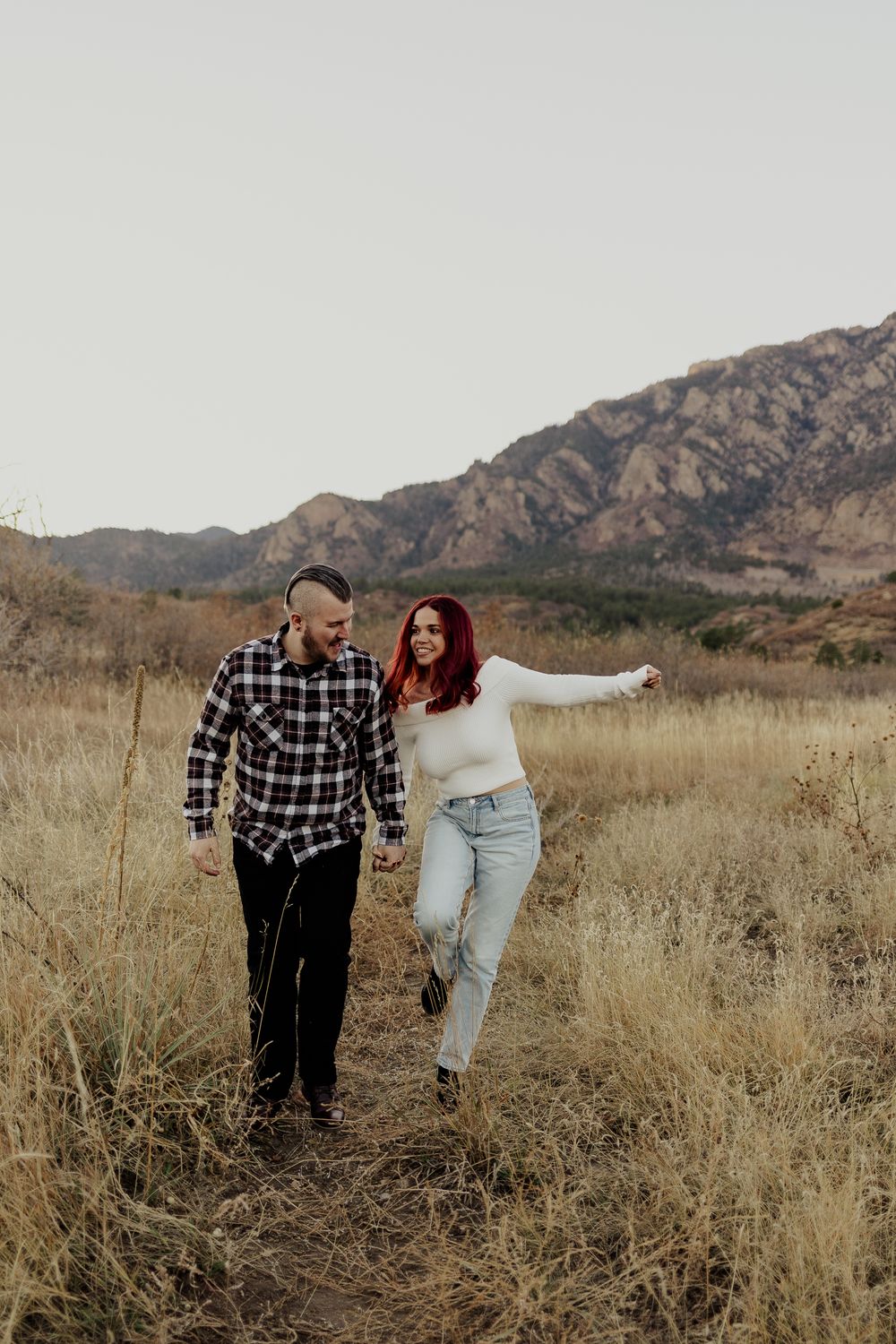 A playful couple dancing together in a golden field with rocky mountains behind them.