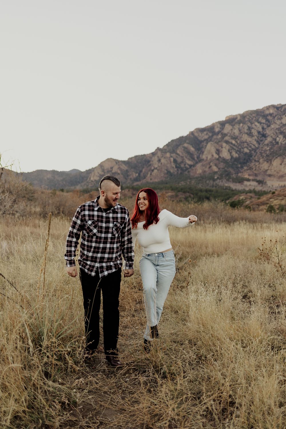 A couple joyfully walks through a field of tall grass with mountains in the background during golden hour.