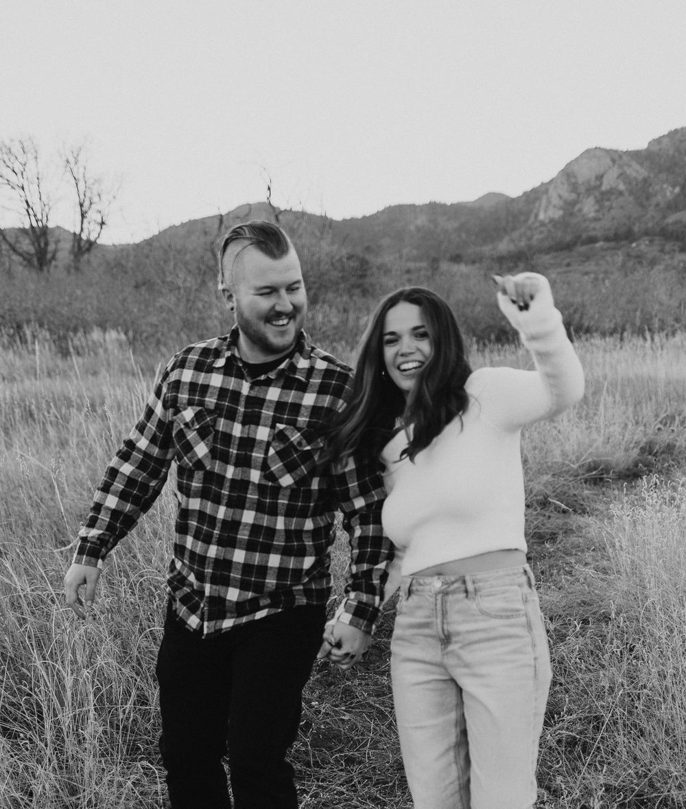 A couple in plaid and white outfits dances playfully together in a field with mountains in the background.