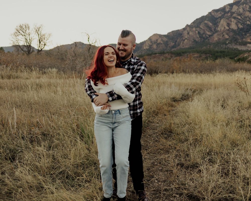 Couple sharing playful moments in a golden field with mountain backdrop during sunset.