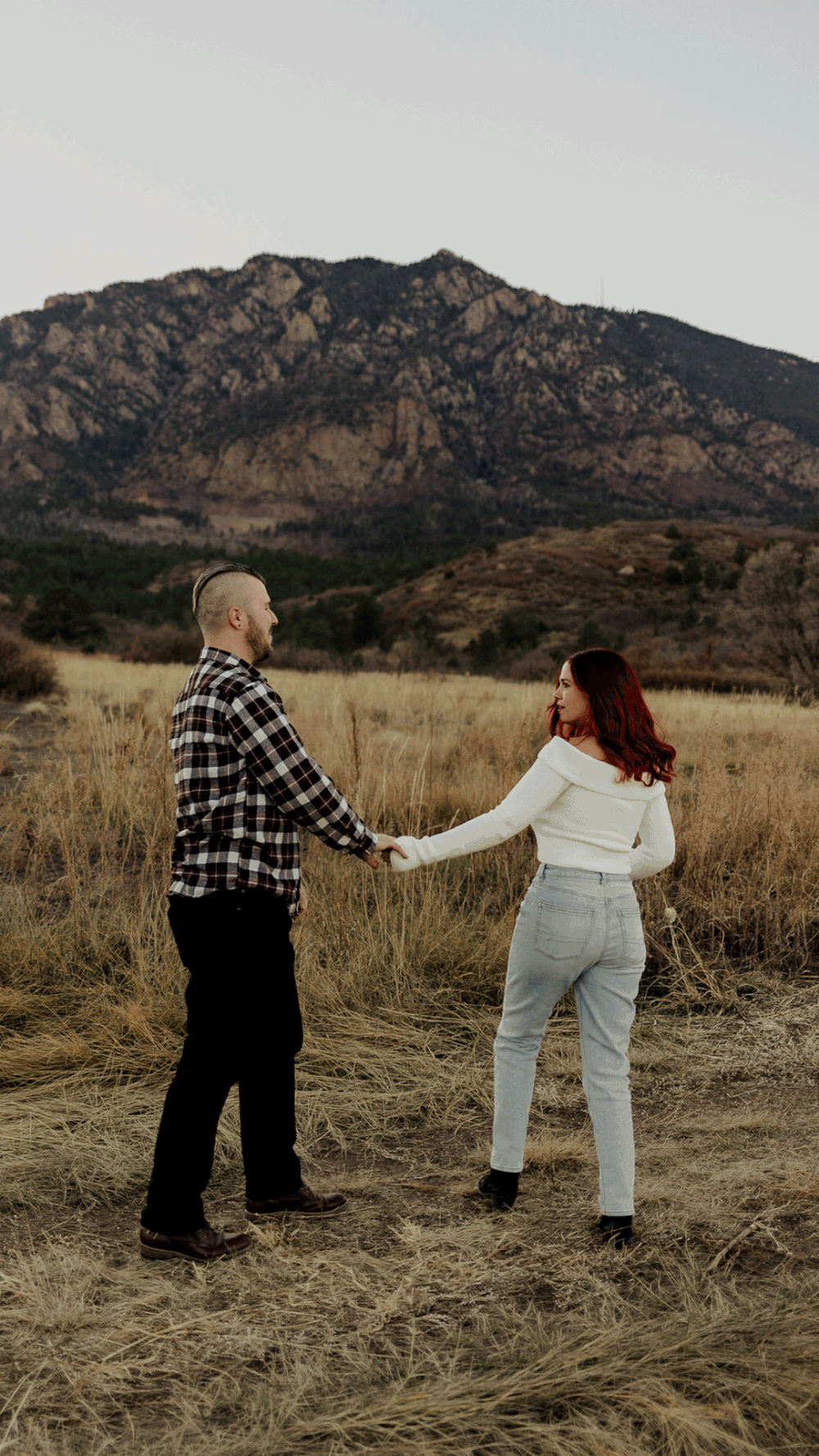 A playful couple dances together in a field with rocky mountains in the background at sunset.
