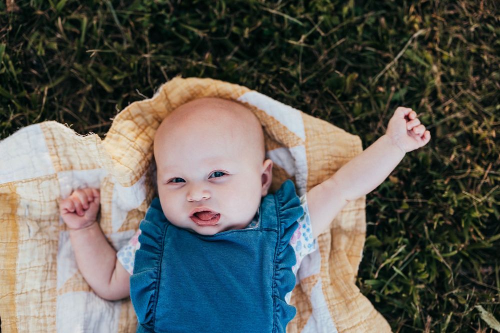 A happy baby in a blue outfit lying on a yellow blanket outdoors on grass with arms stretched out.