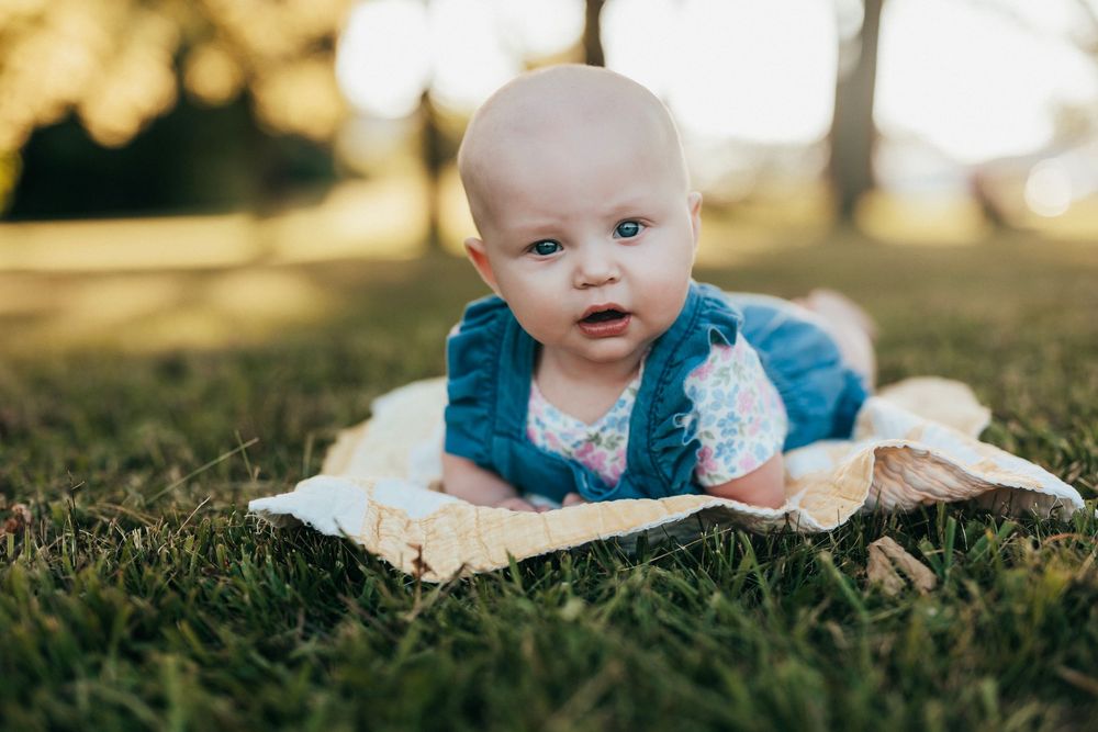 A small baby wearing a blue dress lays in the grass during a sunny outdoor photo session.