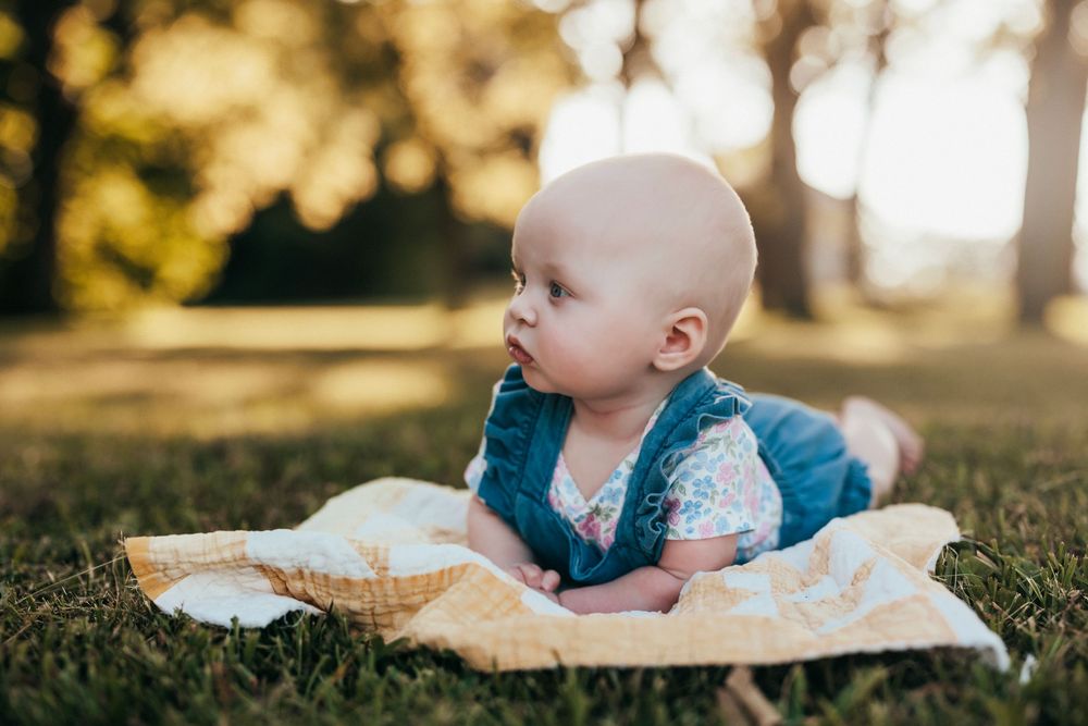 Baby in casual clothing lies on a white blanket in the grass during a sunny outdoor photo session.