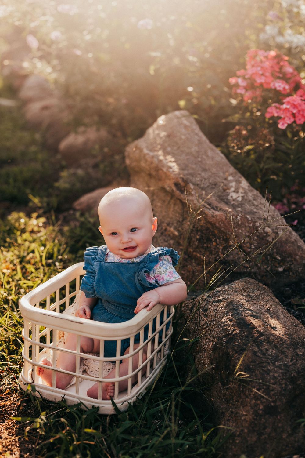 A baby in blue dress sits in white plastic laundry basket against a backdrop of rocks and blooming pink flowers in nature.