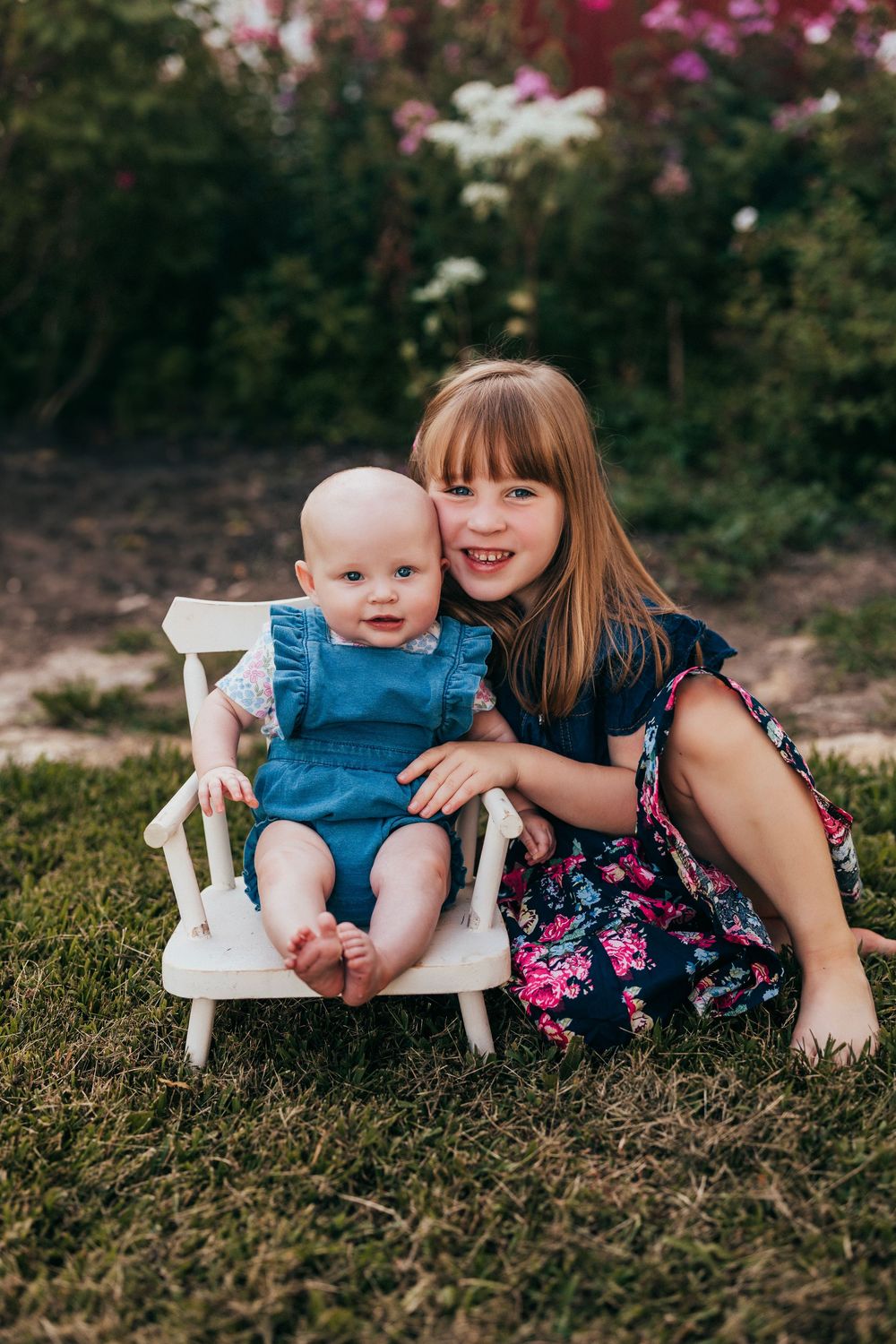 A playful outdoor scene with a baby in a denim outfit seated in a small white chair on grassy ground with floral backdrop.