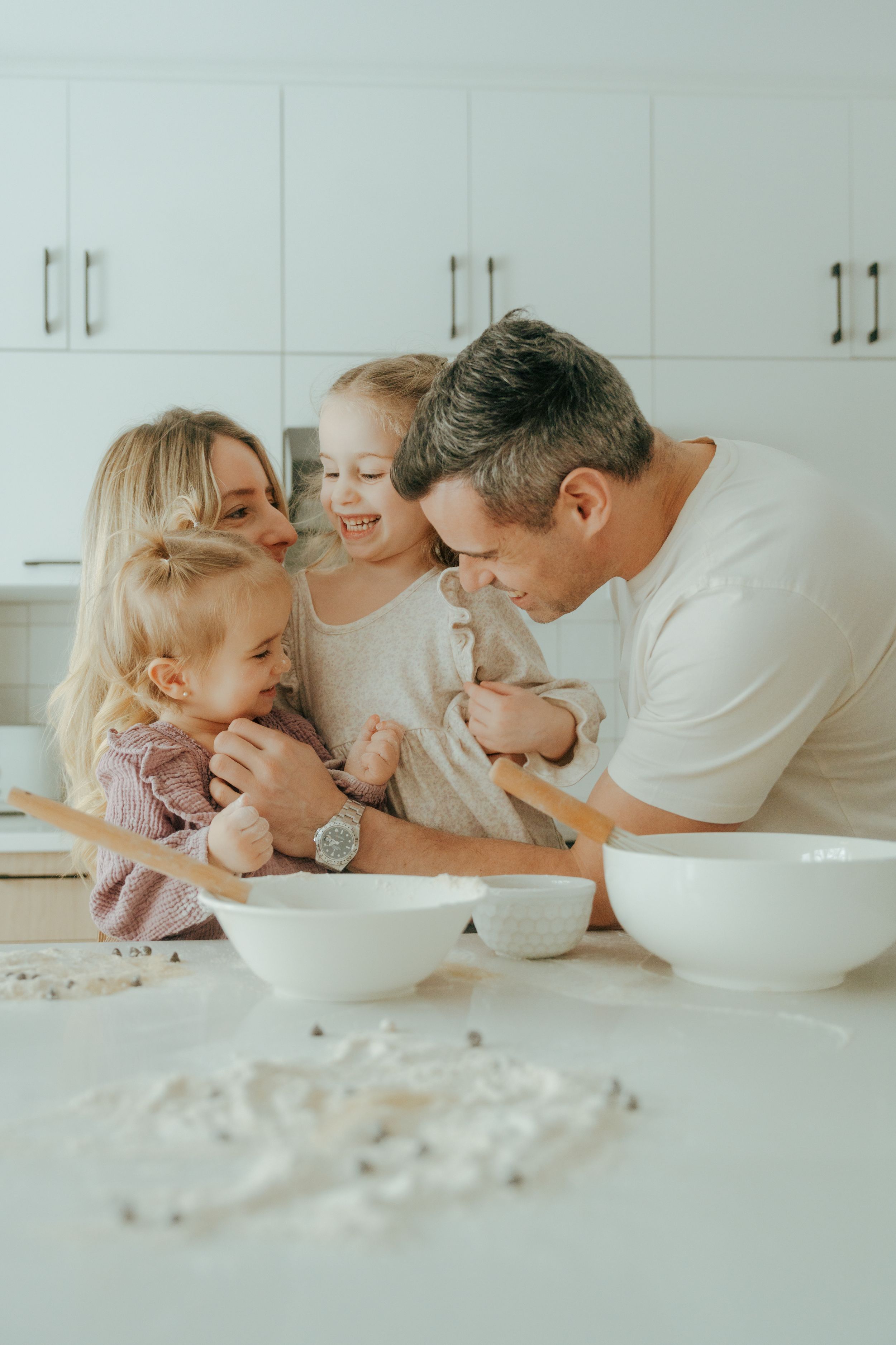 A family enjoys playful moments together while laying on white bedding in a bright, minimalist setting.