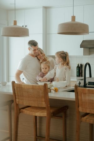 Family gathering in a bright modern kitchen around wooden dining chairs for a casual meal together.