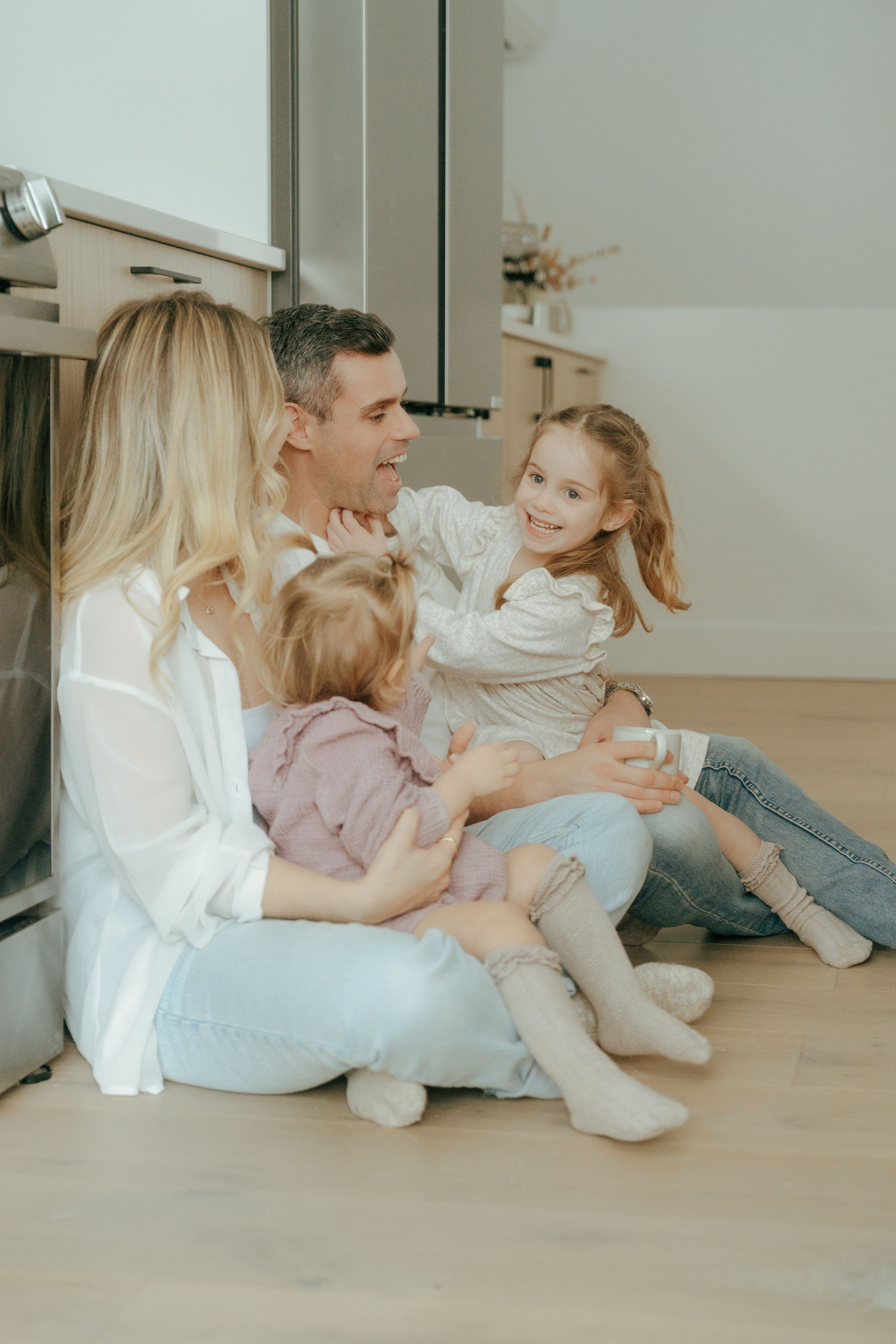 A family sits together on a light wooden floor in a bright room wearing casual white and denim outfits.