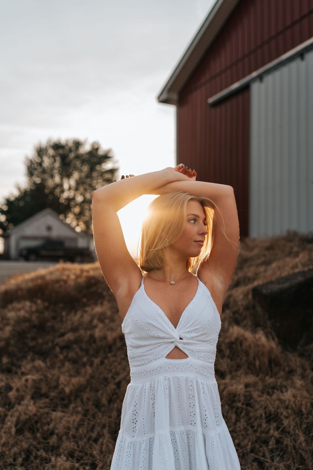 Person in white dress posing in front of a red barn during sunset.