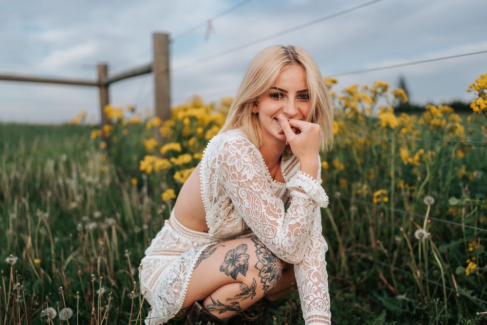 Woman in white lace dress sitting among yellow wildflowers by rustic fence.