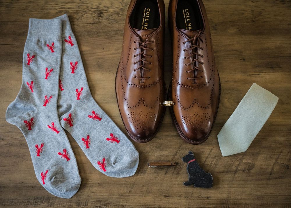 Wedding accessories including brown leather dress shoes, patterned socks, and a light colored tie on wooden surface.