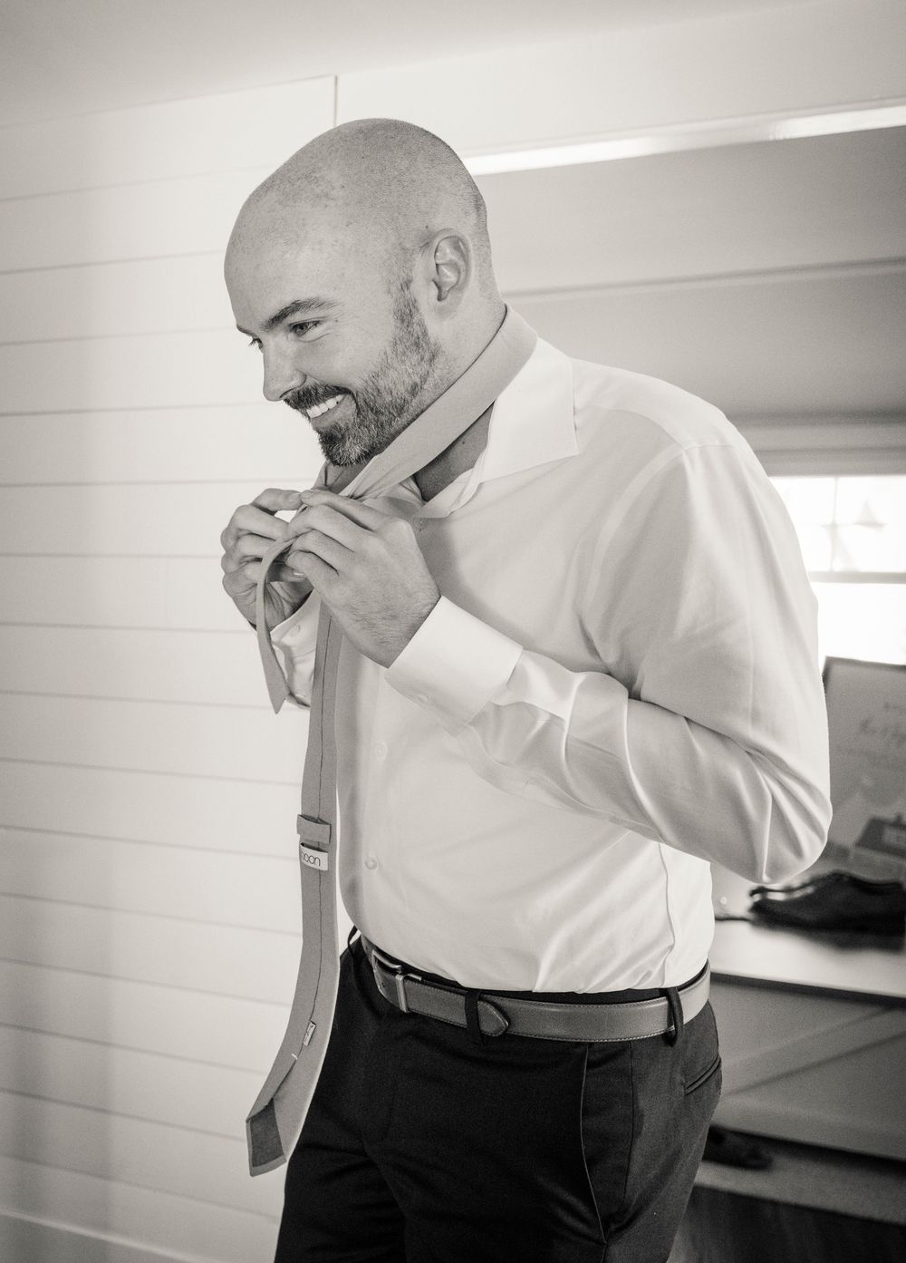 A person adjusts their tie while getting ready in a bright room wearing a white dress shirt.