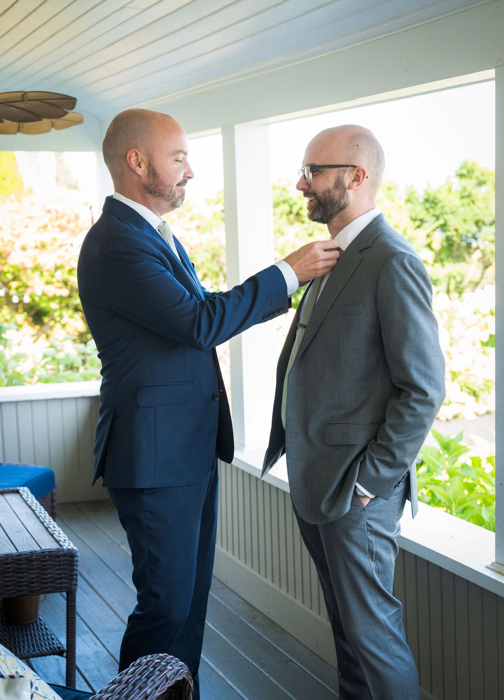 Two people in navy and gray suits adjust each other's ties on a bright porch.
