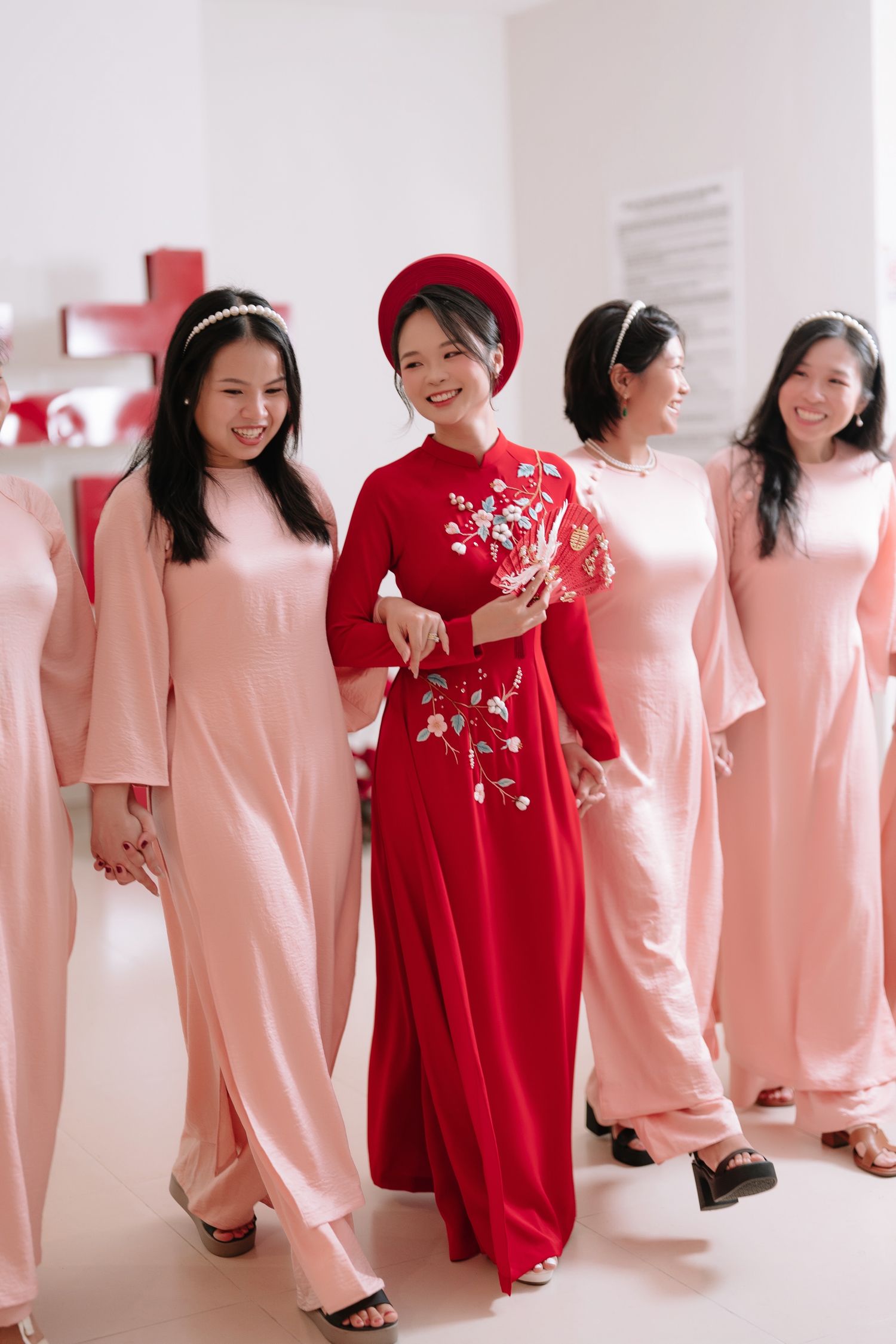 Smiling Vietnamese bride in red ao dai walks with bridesmaids in pink ao dai holding floral fan.