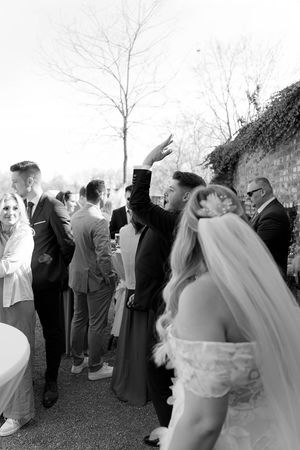 Wedding guests gather outdoors in formal attire as the bride moves through the crowd.