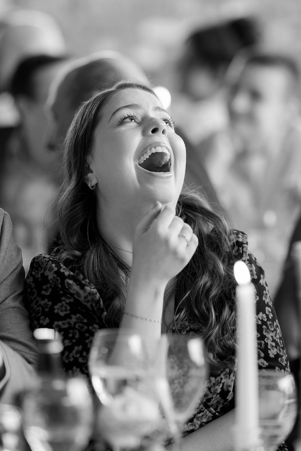 A candid black and white photo of someone laughing joyfully at a dinner table with wine glasses visible.