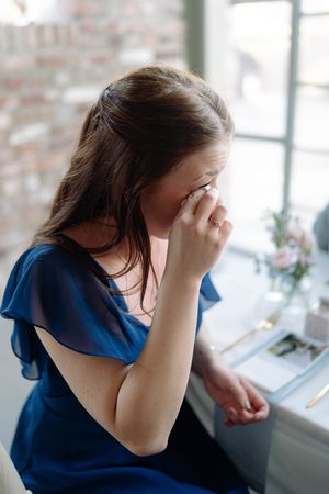 Guest in blue dress shows emotional reaction during wedding reception by window.