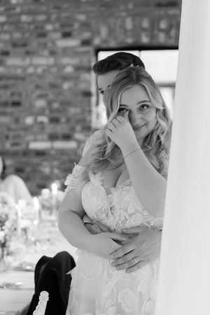 A black and white portrait of someone in a white lace wedding dress peeking around a column.