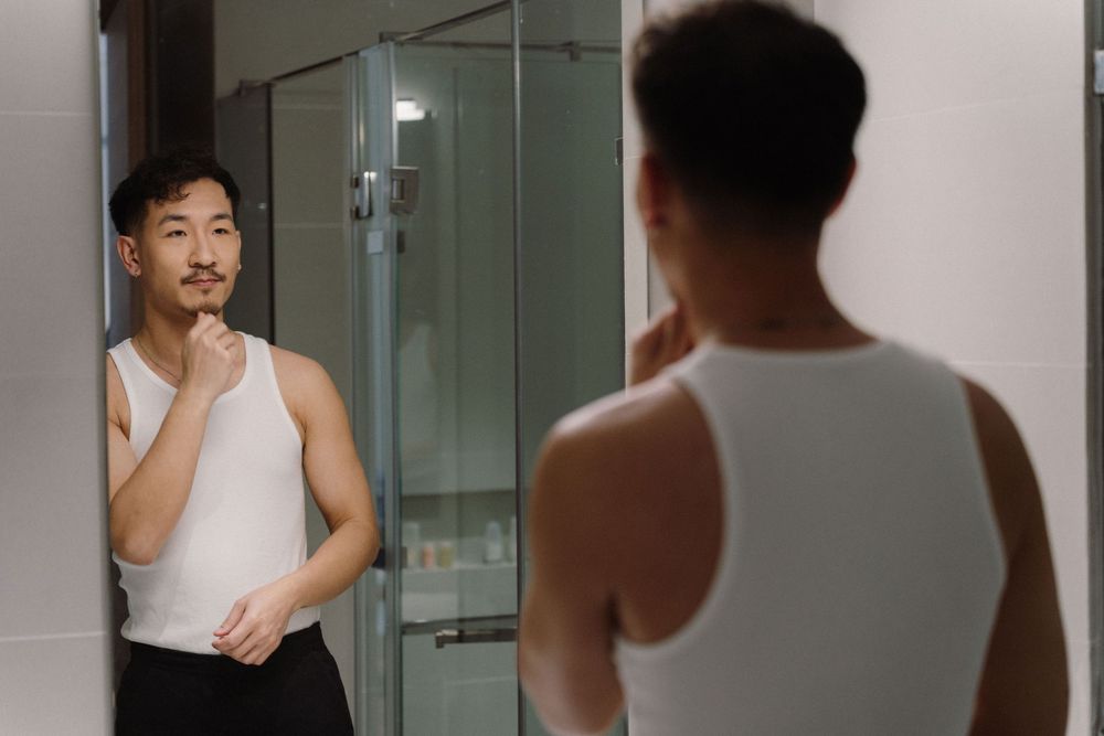 A person in a white tank top looks at their reflection in a bathroom mirror.