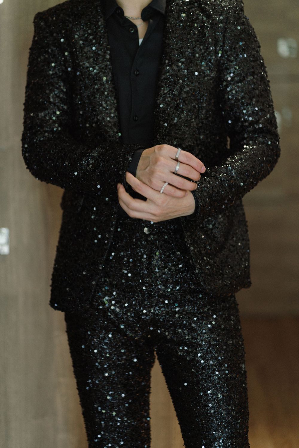 A person wearing a sparkly black sequined pantsuit with glitter details stands indoors.