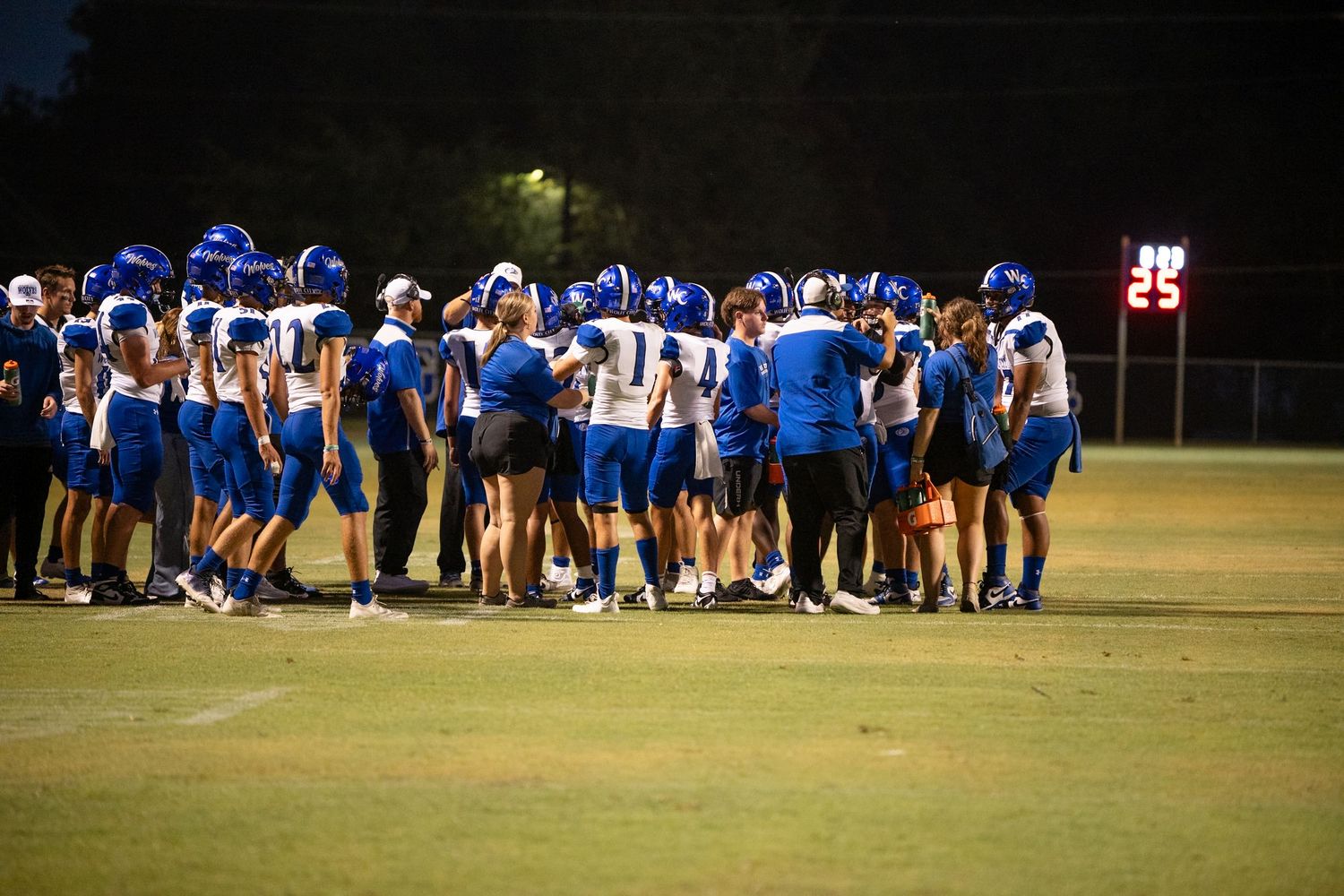 Football teams gather in huddles on a dark field during evening practice session under stadium lights.