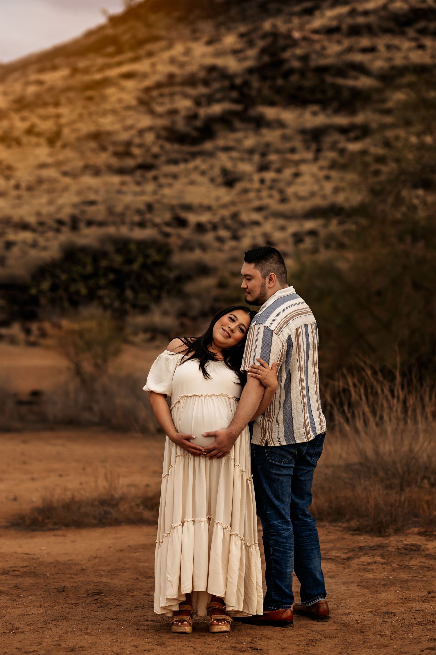 A expecting couple embraces in a romantic sunset photoshoot against a mountainous desert backdrop.