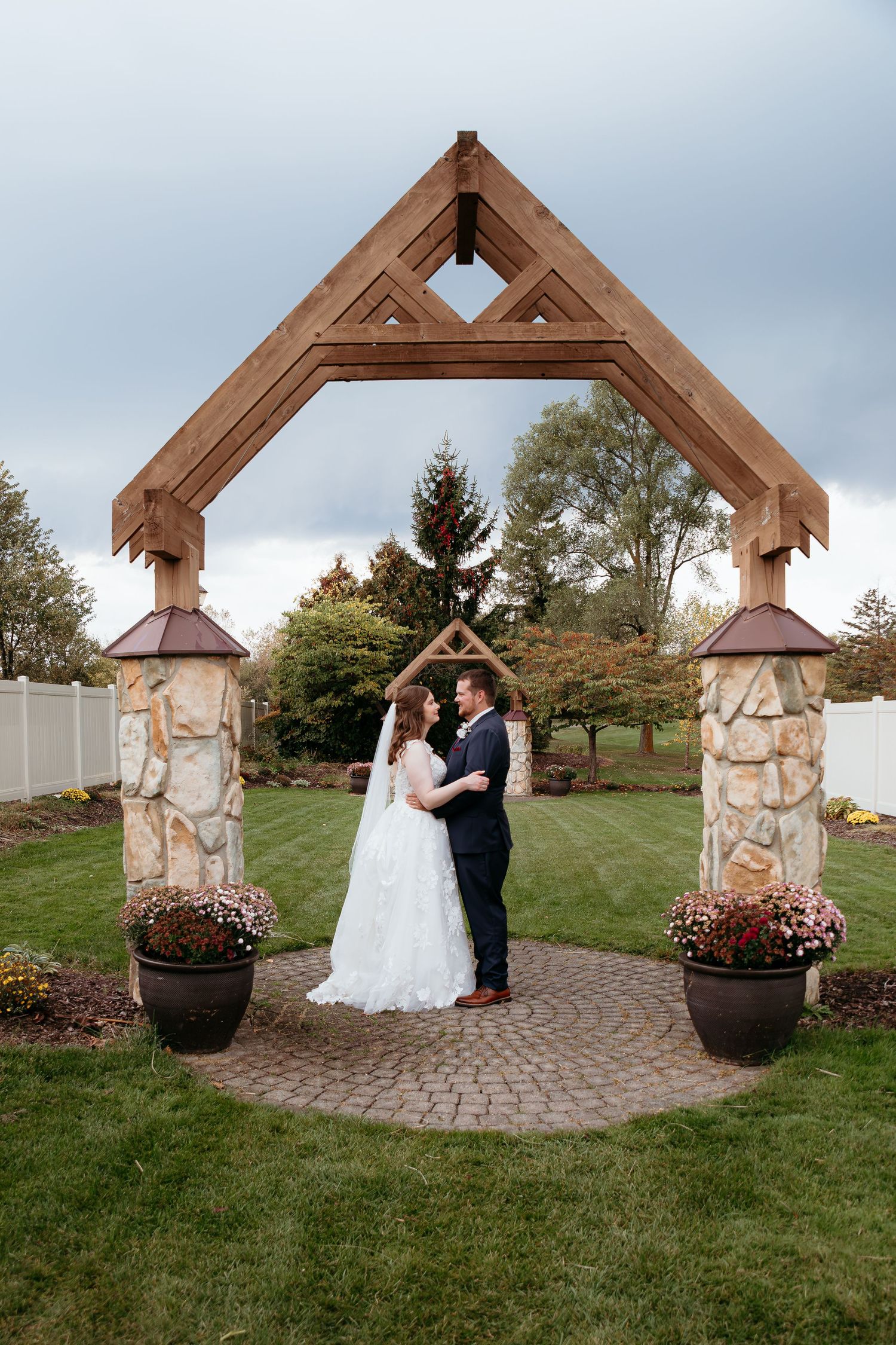 A wedding ceremony under a rustic wooden arch with stone pillars and potted flowers. Glen Oaks Golf course in Farmington Hills, MI