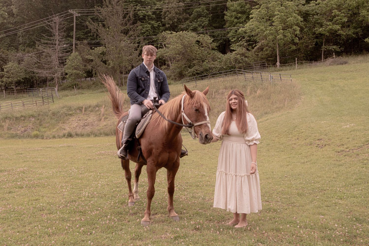 A couple poses in an outdoor field with a brown horse at sunset.