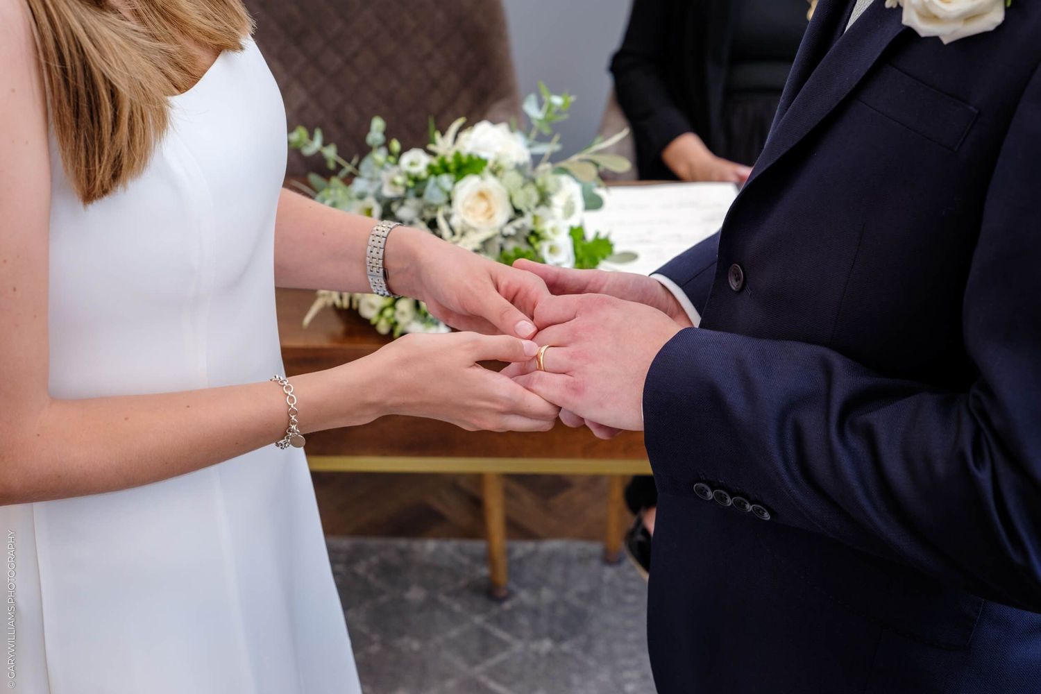 A closeup of exchanging wedding rings during a ceremony with a white dress and navy suit visible.