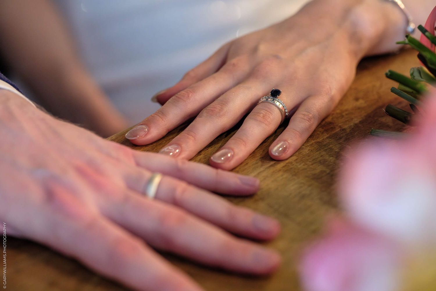 Multiple hands showing wedding rings against wooden backdrop with pink floral decorations.