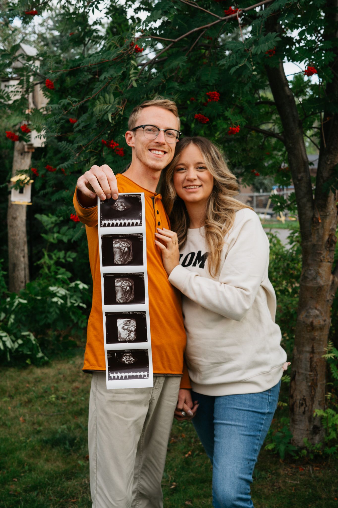 Couple in autumn setting holding ultrasound photos showing their pregnancy announcement in nature.