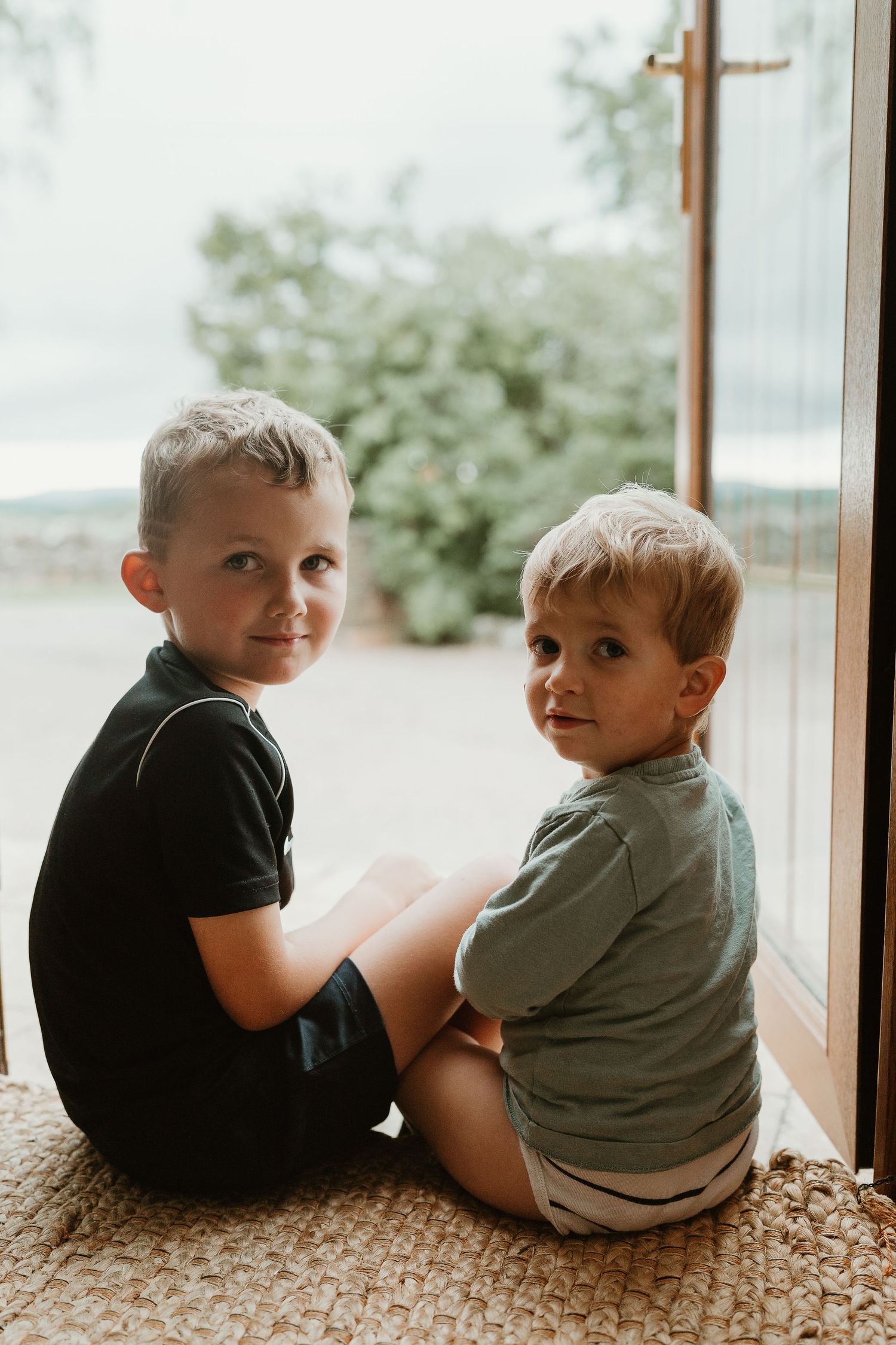 Two young children sit side by side looking out large window in casual indoor setting.