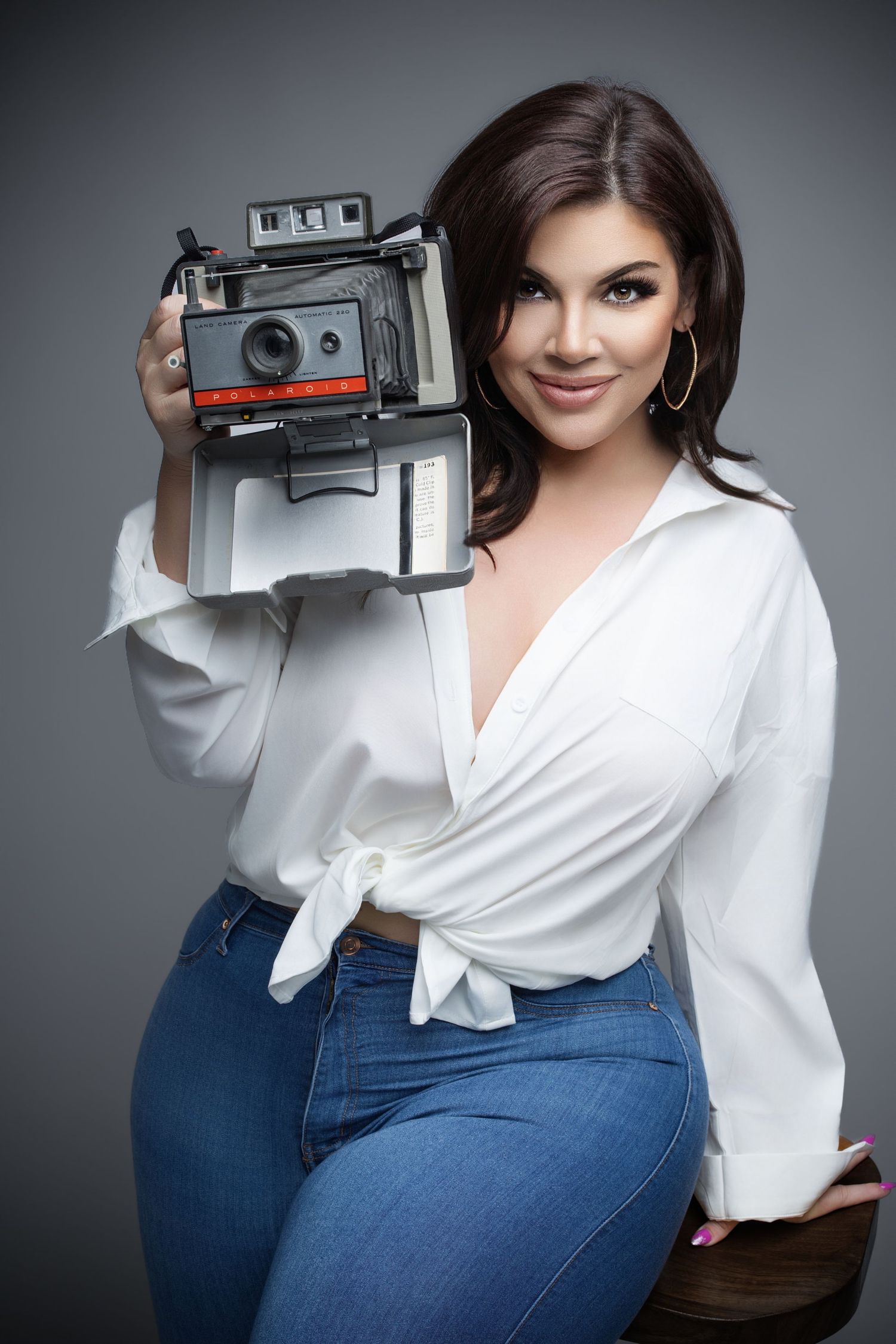 Woman in white blouse and jeans holding a vintage Polaroid camera, smiling in a studio portrait.