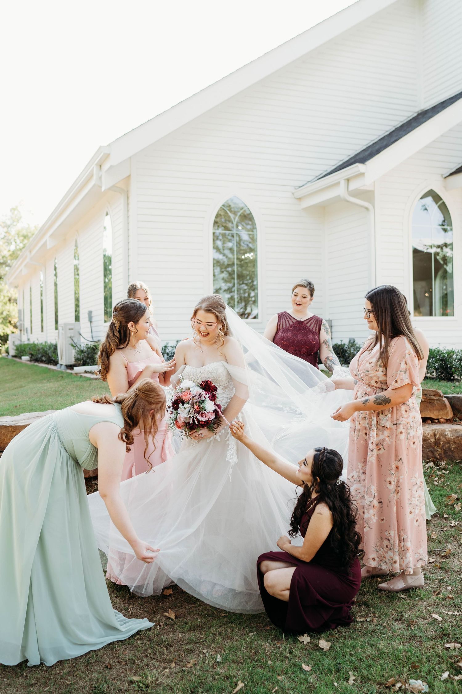 A playful sequence of bridesmaids in pink and mint dresses helping adjust a wedding gown outdoors by a white church.