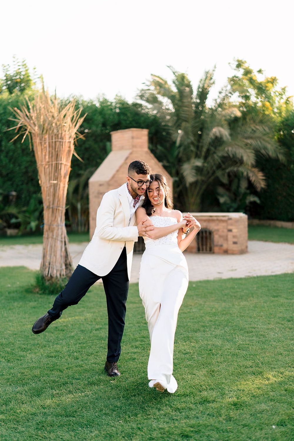 A couple in wedding attire dancing together playfully on a green lawn in front of palm trees at sunset.